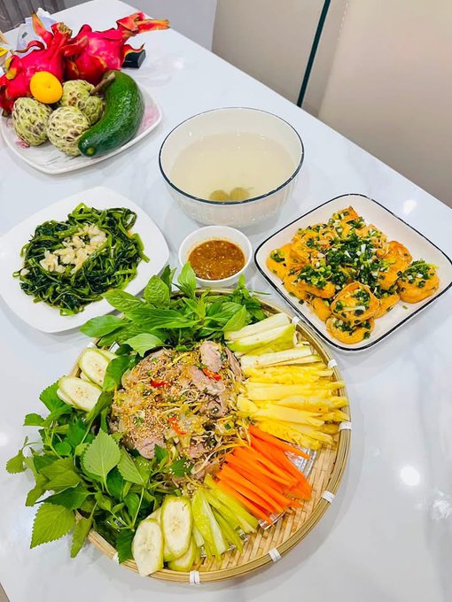A Vietnamese meal spread features a central dish of meat salad surrounded by fresh herbs, cucumber, pineapple, and carrots, alongside a bowl of soup, fried eggs with green onions, sautéed greens, and a plate of tropical fruits.
