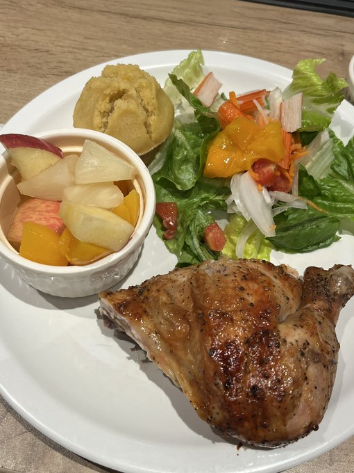 A white plate containing a half roasted chicken, a small bowl of fresh fruit including pineapple and apple slices, a scoop of mashed potatoes, and a mixed green salad with tomatoes and onions.