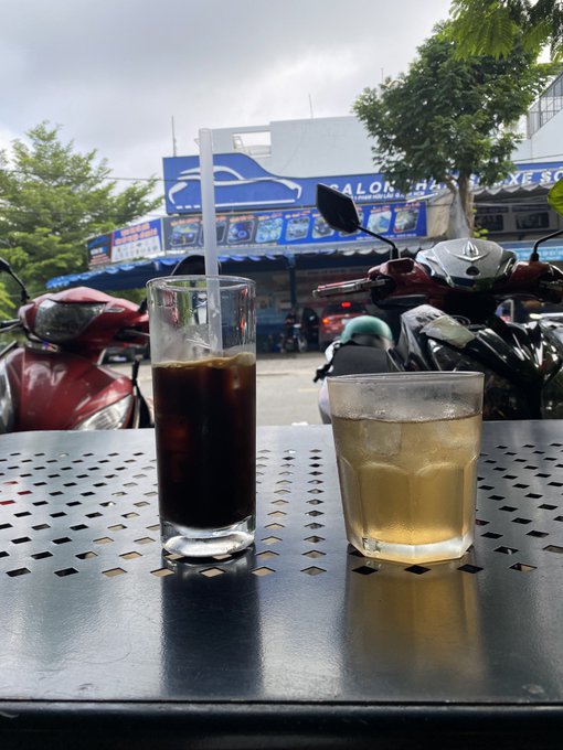 Two glasses of iced drinks—one dark iced coffee and one lighter iced tea—sitting on a perforated metal table at an outdoor café, with parked motorbikes and a salon shopfront in the background.