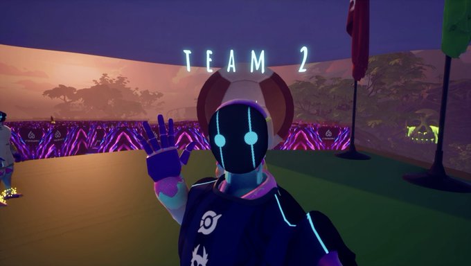 A screenshot from a video game showing a character in a futuristic suit with glowing blue eyes waving, labeled 'TEAM 2' in the background at what appears to be a sports venue.