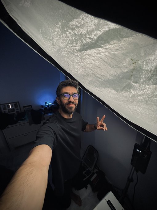 A bearded man wearing glasses takes a selfie in a dimly lit room with blue accent lighting, making a peace sign under a large softbox light.