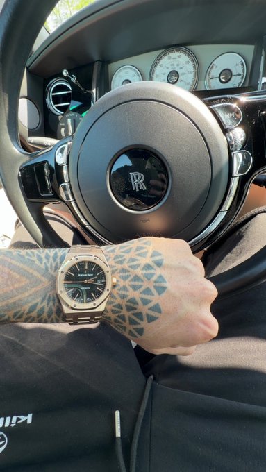 A tattooed hand wearing a luxury watch (Audemars Piguet style) rests on the steering wheel of a Rolls-Royce, with the 'RR' logo visible in the center.