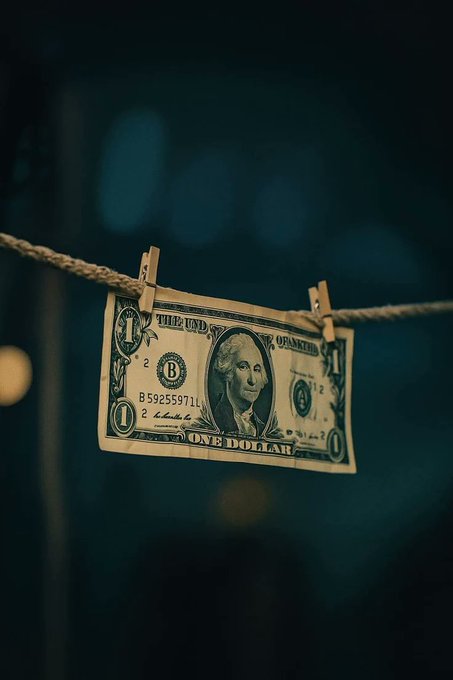 A US one-dollar bill hanging from a rope by clothespins against a dark, blurred background.