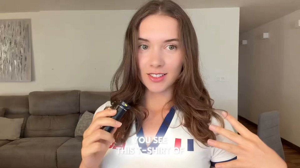 The image features a woman with long brown hair wearing a white shirt that says "You see this? This shirt of mine". She has her hands behind her back, holding a black device or electronic gadget. The background shows a couch against a wall, indicating she is in a living room setting. The post includes text about France and the World Cup, suggesting it might be related to an event or discussion involving these topics.