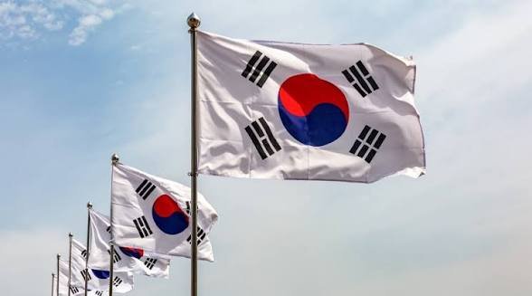 Photograph of multiple South Korean flags (Taegukgi) waving against a partly cloudy blue sky.