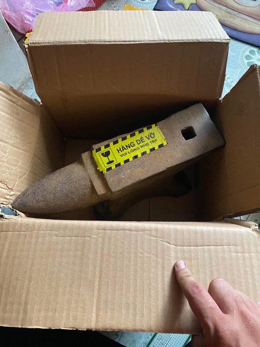 A cardboard box contains a rusty unexploded bomb marked with a yellow warning label reading 'HÀNG DỄ VỠ VUI LÒNG NHẸ TAY' (Fragile items, please handle gently), with a finger pointing at it.