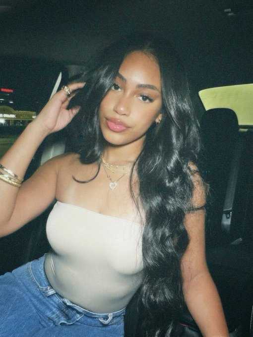 A woman with long wavy black hair takes a selfie in a car at night, wearing a cream-colored tube top and denim, with layered gold necklaces and rings.