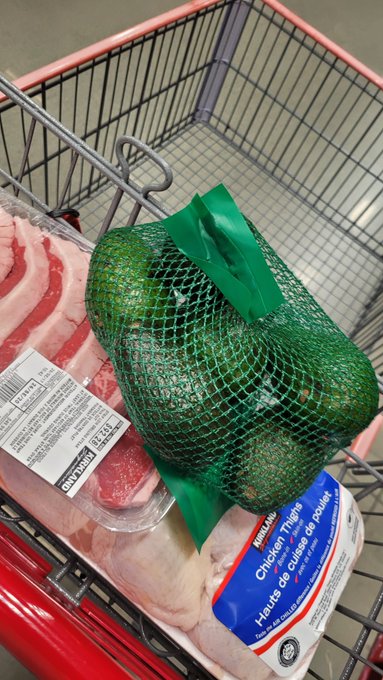 A shopping cart contains raw pork, a bag of avocados in green mesh, and Kirkland chicken thighs on the bottom.