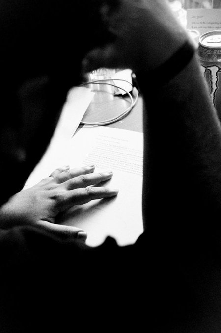 A black-and-white close-up shows a hand resting on a script as a figure wearing headphones leans over it, with a Monster energy drink and a small card visible in the background—capturing an intimate moment of speech preparation in a Prague hotel room.