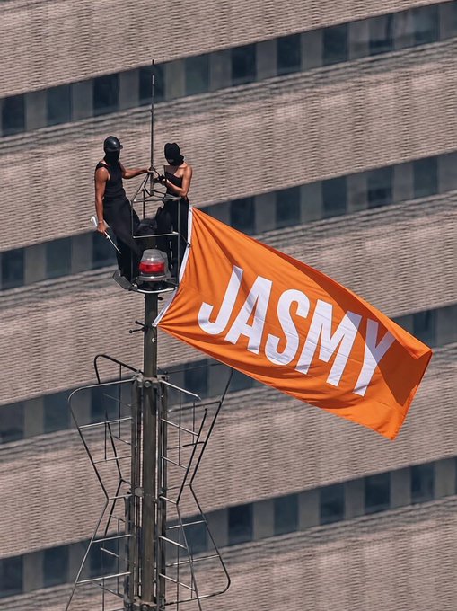 Two workers in a basket on a tall pole raising an orange 'JASMY' flag against a city building backdrop.