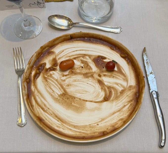 A bowl of coffee or cappuccino with decorative latte art and two small fruits (a cherry tomato and a cherry) arranged to look like a face, served with a fork, knife, and spoon on a table.