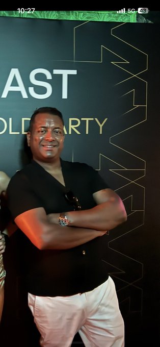 A man in a black polo shirt and white pants stands smiling with his arms crossed in front of a backdrop with gold lettering, including the visible text 'AST' and 'OLD PARTY'.
