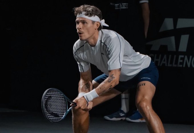 A focused tennis player wearing a white headband and gray shirt crouches in ready stance, gripping a tennis racket as he prepares to receive a serve. The image complements the "$100 → $10,000 Ladder Challenge" post by visually capturing the competitive intensity and strategic anticipation required in any high-stakes climb toward a goal.