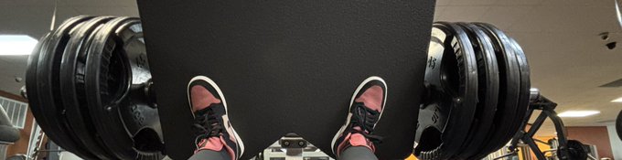 A first-person perspective photo looking down at a pair of pink and black sneakers on a gym leg press machine loaded with heavy weight plates.