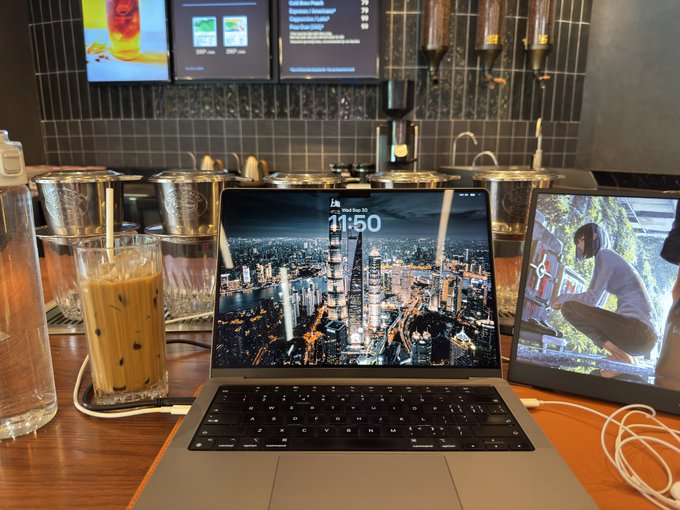 A laptop and tablet sit on a wooden table in a cafe setting next to an iced coffee drink, with the laptop screen displaying the date and a cityscape wallpaper.
