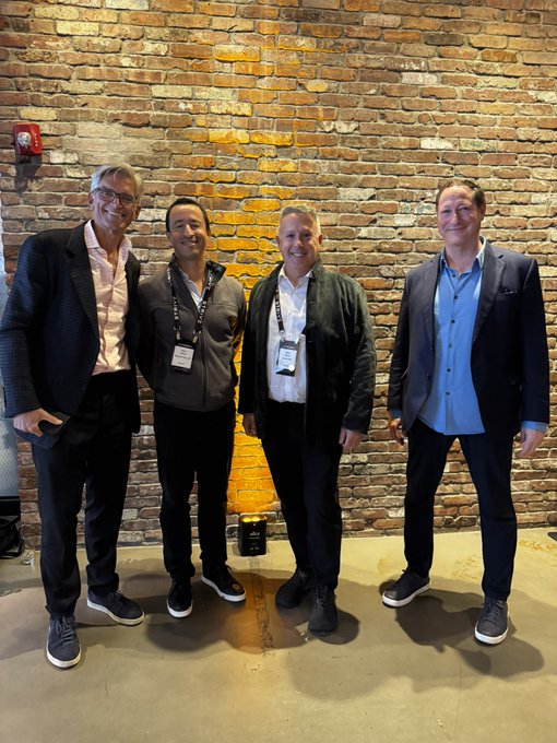 Four men in business attire pose together at what appears to be a conference or networking event, standing against an exposed brick wall with warm accent lighting.