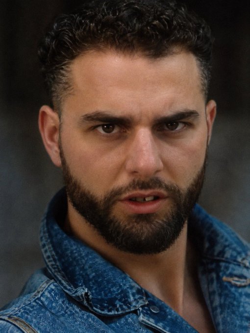 A close-up portrait of a young man with dark curly hair and a full beard, wearing a blue denim jacket against a gray background.
