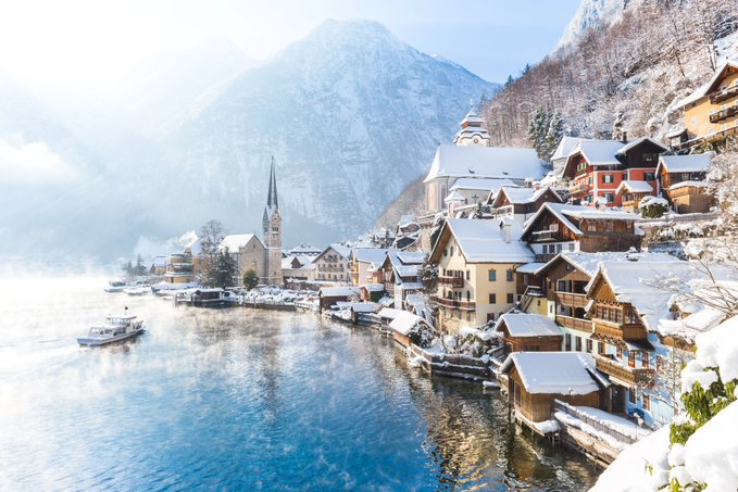 A scenic winter photograph of a snow-covered lakeside village with a prominent church spire, resembling Hallstatt, Austria.