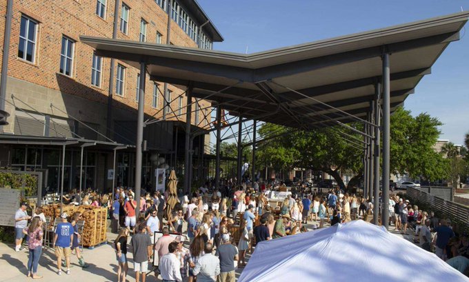 A large crowd of people attending an outdoor market or festival under a metal canopy structure on a sunny day.