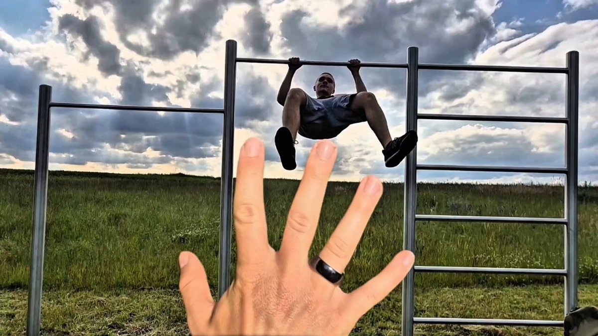 The image features a man hanging from a bar with his hand outstretched, wearing black shoes and sunglasses. The background shows a cloudy sky over a grassy field. There are two gray metal bars visible in the scene, one of which has a ring attached to it. A caption on the image reads "Finally a hardware wallet that fits my lifestyle."