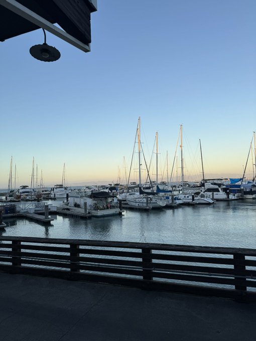 A marina is shown, featuring numerous sailboats docked in calm waters against a clear sky, viewed from a deck with a wooden fence.

The accompanying post "GM. Last day of vacation. Happy Monday everyone." suggests the user is sharing a peaceful morning view from their final day of vacation at this harbor, bidding good morning to their followers.