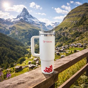 A white TRON-branded tumbler with a handle and straw sits on a wooden railing in front of the iconic Matterhorn mountain and a Swiss alpine village. The image pairs with the caption to visually evoke TRON's expansion into real-world infrastructure—mirroring the post's theme of TRON-native payment rails powering decentralized GPU compute marketplaces for AI workloads.