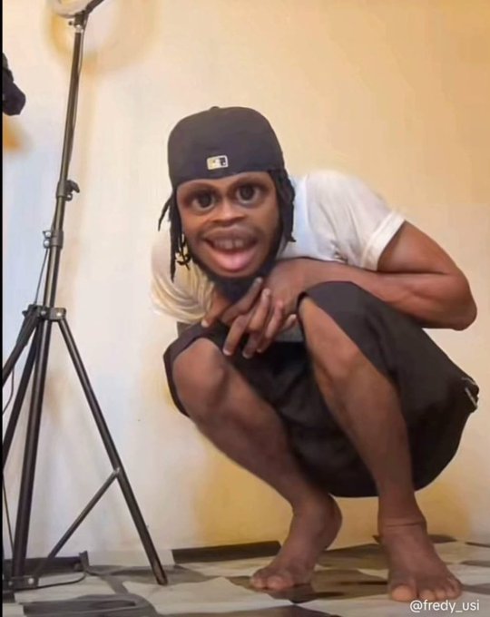 A person wearing a black cap with a face filter applied is squatting indoors next to a studio light stand, with the watermark '@fredy_usi' in the corner.