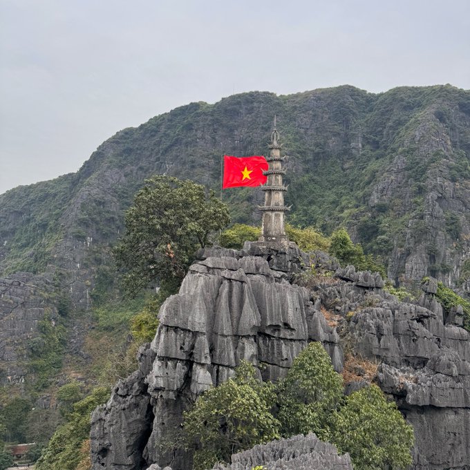 A photograph shows a Vietnamese flag flying next to an old stone tower perched atop a rugged, rocky mountain formation surrounded by green vegetation.