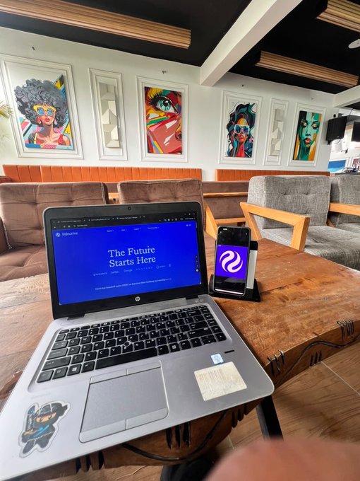 The image shows a laptop computer sitting on top of a wooden table, with a cell phone placed next to it. The background features several paintings hanging on the wall above the table. A caption below the image reads "Happy weekend Ninjas Spend less time looking at the $INJ chart today and touch grass. It's weekend". This suggests that someone is using their laptop for work or leisure, while also enjoying some downtime by touching the grass outside.