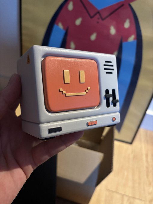 A hand holds a small retro-style TV figurine featuring an orange screen with a pixelated smiley face, set against a blurred background with a red and blue logo.
