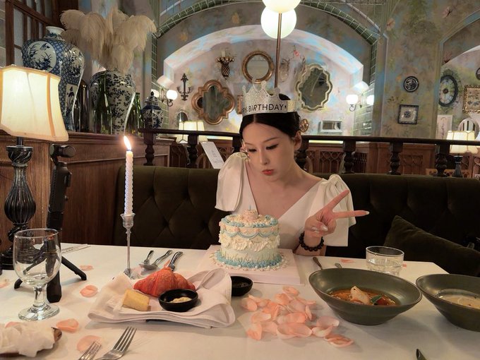 A woman in a white dress blows out a candle on a small blue and white birthday cake while wearing a "BIRTHDAY" tiara at an elegantly decorated restaurant. The caption "Good morning, leveling up from the very first day of September!" suggests she is celebrating her September birthday.