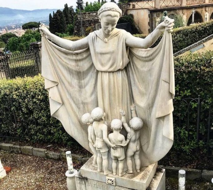 A stone statue of a woman holding a flowing cloak over several children, with 'SILVIA MARININI' inscribed on the base, set in a garden with a scenic hillside town view in the background.