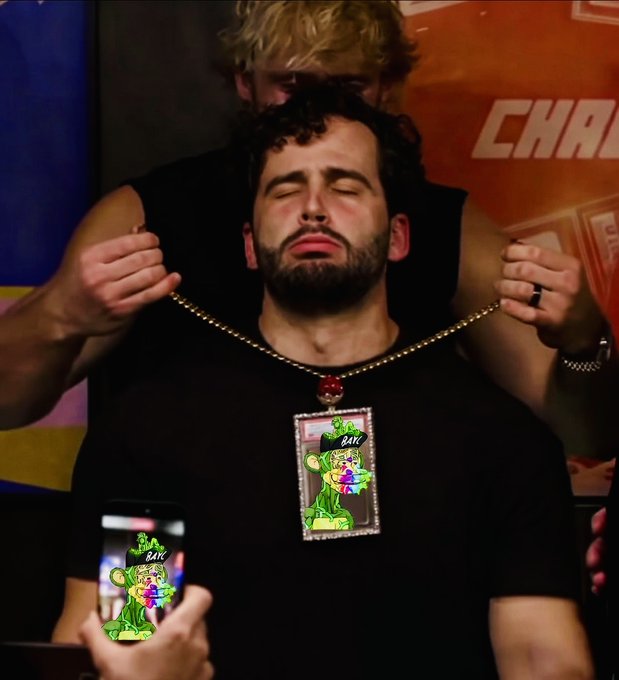 A man with a beard wearing a black t-shirt with eyes closed as another person places a large bejeweled pendant necklace featuring a green cartoon character with a baseball cap over his head.