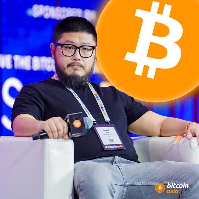 The image features an Asian man sitting in a chair, wearing glasses and holding a microphone. The background of the image shows a large Bitcoin logo with the text "Bitcoin" written on it. The caption below the image reads "Focus on what is in the image and how it relates to the post".