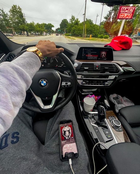 A driver sits behind the wheel of a BMW with a Playboi Carti album ("Whole Lotta Red") playing on the car's infotainment screen and on their iPhone, which is connected via cable on the center console. The caption "How much is your Breakfast today?" plays off the early-morning drive vibe, prompting followers to share their Solana wallet addresses in the replies.