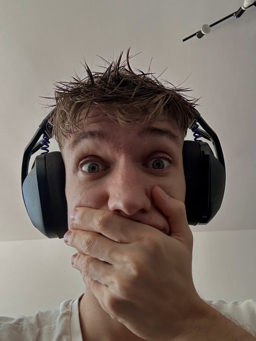A close-up selfie of a young man with messy hair wearing large black and blue headphones, looking surprised with wide eyes and his hand covering his mouth.