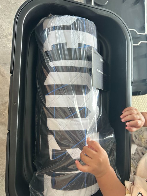 A child unwraps a Gate-branded black and white camping mat set still in its plastic packaging inside a black case. The caption aligns with the post celebrating the delivery of Gate's giveaway camping gear in the described color scheme.