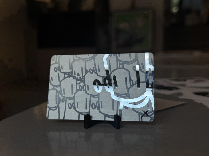 A small rectangular canvas or card featuring grey cartoon-style artwork is displayed on a black stand, with more artwork visible on the surface behind it.