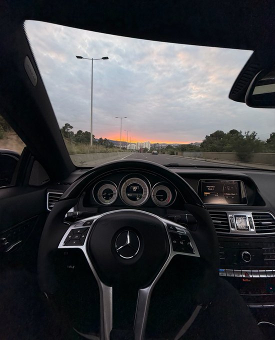 View from inside a Mercedes-Benz driving on a highway at sunset, with the iconic three-pointed star logo visible on the steering wheel and city skyline in the distance.