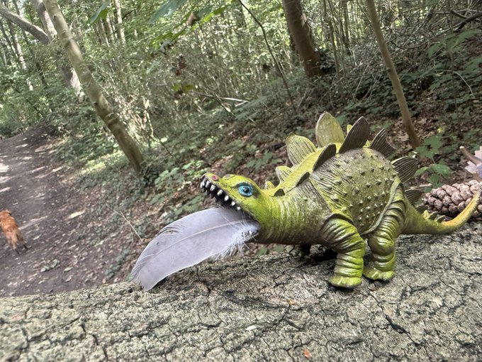 A green plastic toy stegosaurus with a white feather in its mouth sits on a tree branch in a wooded area.