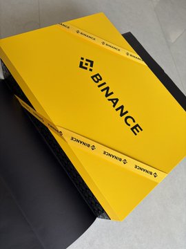 The image features a yellow box with a black ribbon, which has "Binance" written on it. The box appears to be a gift or present for someone named Binance. The background of the image is white, making the colors of the box stand out even more.