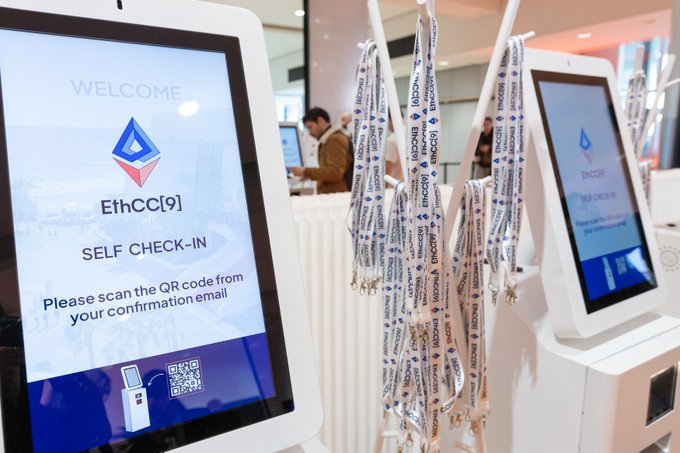 Self check-in kiosks at an EthCC[9] conference display welcome screens asking attendees to scan QR codes, with branded lanyards hanging nearby.
