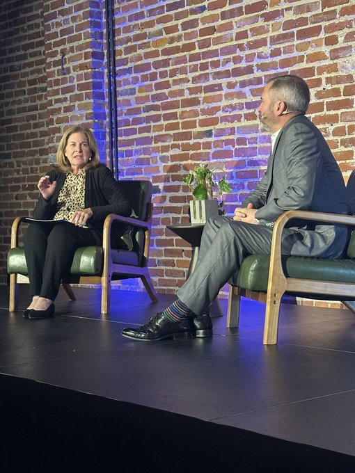 Two people sit in green chairs on a stage against a brick wall, with a woman on the left gesturing while speaking to a bearded man in a suit on the right, alongside a small table with a vase of flowers. The setting appears to be a panel discussion at HDAX, consistent with the post describing a conversation about XRP and Ripple's legal clarity following a court ruling.
