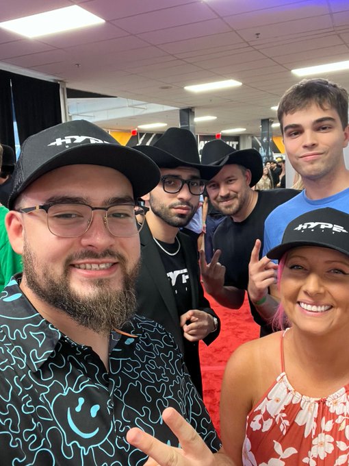 A selfie of five people posing together at an event, with some wearing 'HYPE' branded caps and others in cowboy hats, taken on a red carpet area.