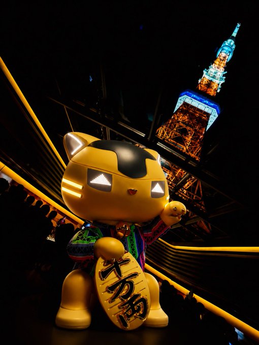 A glowing robotic MANEKINEKO (lucky cat) figurine stands prominently in the foreground at a nighttime event, with Tokyo Tower illuminated in orange behind it. The cat holds a gold coin inscribed with "千万両" (ten million ryō), symbolizing prosperity, fitting the post's theme of celebrating the mascot as a cherished symbol of community and future growth toward Mainnet and Breed.