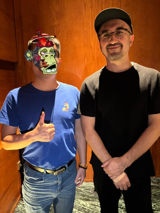 Two men stand side by side smiling; the man on the left has his face covered by a colorful cartoon mask and gives a thumbs up while wearing a blue t-shirt, and the man on the right wears a black cap, glasses, and a black shirt.