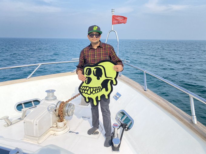 The image features a man standing on a boat with his arms raised above his head while holding up a skull decoration. The man appears to be wearing sunglasses and has a hat on, giving him a cool and relaxed vibe. The background of the photo shows an ocean view, adding to the overall atmosphere of the scene.