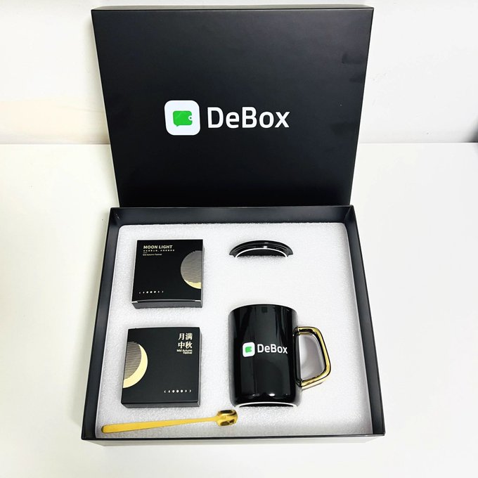A black DeBox branded gift set containing a black mug with gold handle, coffee packages labeled 'MOON LIGHT' and '月满中秋 Mid-Autumn', a lid, and a gold spoon arranged in foam packaging.