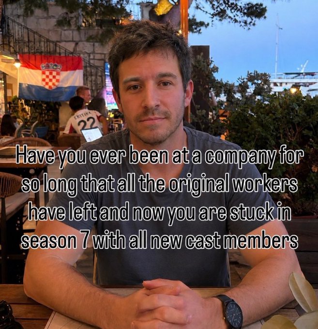 Photo of a man sitting at a restaurant with a Croatian flag visible in the background, overlaid with the caption "Have you ever been at a company for so long that all the original workers have left and now you are stuck in season 7 with all new cast members."