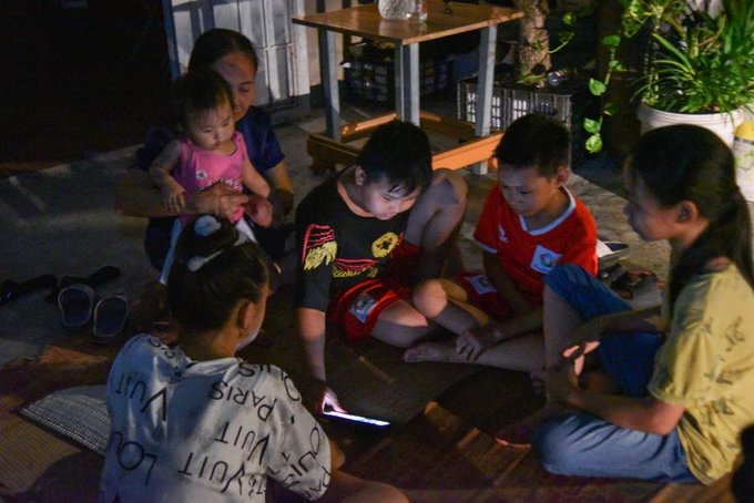 A group of people, including children and adults, sitting together on the floor at night looking at a smartphone screen, with one adult holding a toddler in a pink outfit.